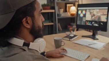 Atkuyruklu ve sakallı genç Orta Doğulu adamın evde kurgu yazılımında fotoğraf ya da görüntü üzerinde çalışırken yakın plan çekimleri ve müzik dinlemek için kablosuz kulaklık takması.