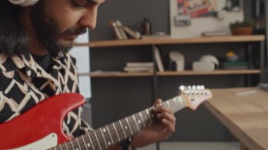 Uzun siyah saçlı, sakallı ve kulaklıklı genç bir adamın evde oturup elektro gitarla müzik çalarkenki yakın çekimi.