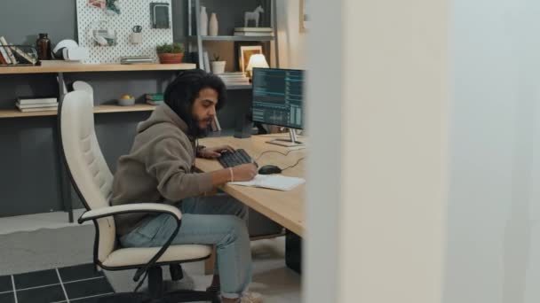 中東の若い男性ソフトウェアエンジニアが自宅のオフィスの椅子に座り 大きなコンピュータスクリーン付きの机に座り キーボードにコードを書き 入力するフルショット — ストック動画