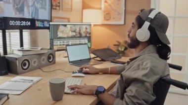 Özürlü genç erkek prodüksiyon tasarımcısının evde, tekerlekli sandalyede oturup müzik dinlerken, kahve içerken ve kurgu yazılımında video klipleri üzerinde çalışırken orta ölçekli bir fotoğrafı.