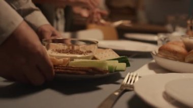 Yahudi aile üyelerinin Hanuka bayramı yemeği için hazırlanan yakın plan fotoğrafları. İsimsiz bir adam humus, salatalık ve kraker tabağı koyuyor ve kız çatal bıçak koyuyor.