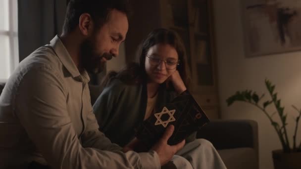 キッパのユダヤ人の父親と幼い娘が家でソファに座って ダビデの星と一緒にトラーの本を保持し 祈りを読み 勉強し 聖書について話し合う中側のショット — ストック動画