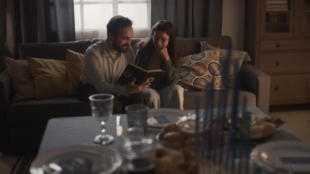 若い女の子とソファーに座って トーラの本を保持し 聖書を読み 議論し ハンカイヤとぼやけた前景でお祝いのテーブルに座ったキッパのひげ付きユダヤ人の男の完全なショット — ストック動画