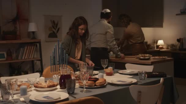 自宅でハヌカの夕食のために準備するユダヤ人家族のミディアムショット テーブルの上にカトラリーを置く娘 伝統的な食べ物とプラッターを運ぶ父親 キッチンでオーブンからオペレルを取る母 — ストック動画