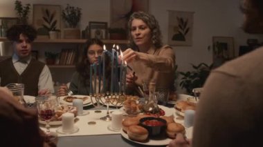 Yahudi annenin Hanukkah yemeği sırasında ailesiyle birlikte masada oturduğu, Hanukkiah 'ta mum yaktığı, oğlunun ve kızının izlediği ve konuklarla sohbet ettiği orta boy bir fotoğraf.