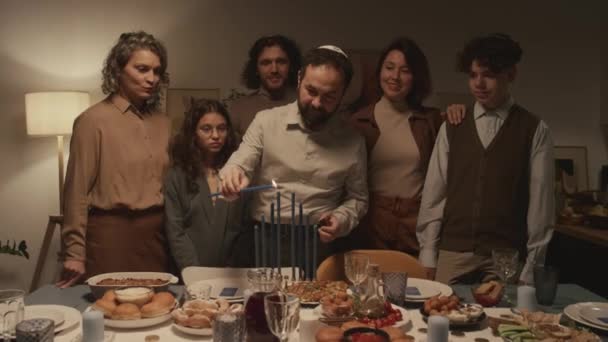 ハヌカのお祝いで夕食テーブルの周りに立っている6人のユダヤ人の家族のミディアムショット キッパの男は 燃えるシャムシを保持し ハンカイヤにろうそくを点灯し 誰もが見ている間 — ストック動画