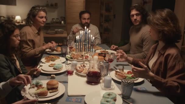 お祝いのテーブルの周りに座っている6人のユダヤ人家族のミディアムショット ハヌカを祝うために夕食を食べる 若い女性はエネルギッシュなジェスチャーで面白い話をし みんなが聞いている — ストック動画