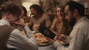 Kippah 'ta Yahudi bir adamın ailesi ve arkadaşlarıyla birlikte Hanuka' da oturmuş kutsal Tevrat kitabından bölümler okurken kapağında Davut 'un yıldızı ve oturup dinleyen insanlar.