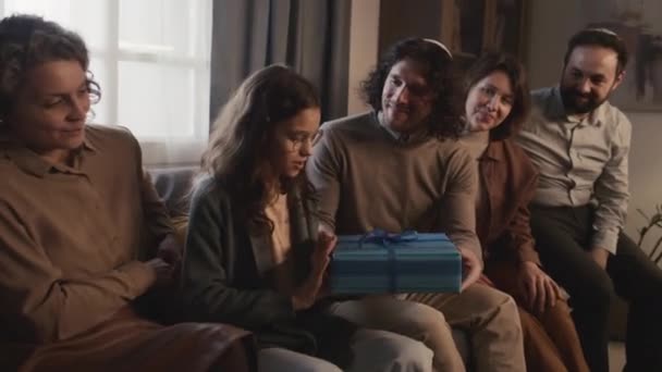 幸せなユダヤ人の家族のミディアムショット キプパの父親 ソファに座って息子と娘 十代の妹ハヌカの贈り物を与える若者 そして 中にあるものを推測するために 少女の揺れる箱 — ストック動画