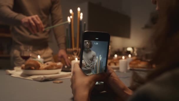 お祝いの食べ物を持ってテーブルに立っている若いユダヤ人の中型ショット ハンカイヤにろうそくを注意深く点灯し スマートフォンを持って彼を撮影する若い女性の手 — ストック動画