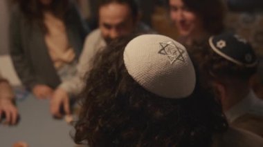 Yahudi geleneksel başlığının yakın plan seçmeli fotoğrafı, Kipah, Davut 'un yıldızıyla süslenmiş, Hanuka partisinde ailesi ve arkadaşlarıyla dreidel oyunu oynayan genç bir erkeğin başında.