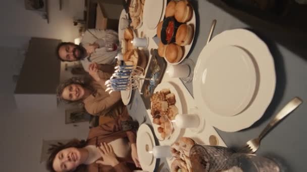 ハヌカのお祝いのテーブルの周りに座っている家族のビデオを作るスマートフォンを持つ若いユダヤ人の少女の垂直のセルフィーショット カメラで笑顔と振動 — ストック動画