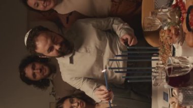 Orta yaşlı sakallı Yahudi adamın Hanuka yemekleriyle bayram masasında durup şampuanla Hanukkiah 'ı yaktığı ve neşeli çocukların izlediği orta boy dikey bir fotoğraf.