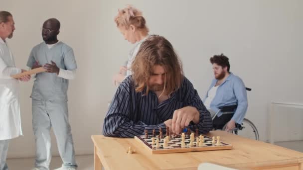 精神病院のレクリエーションルームのテーブルに座ってチェスを演じる若い無意識の認知症に苦しんでいる白人男性 — ストック動画