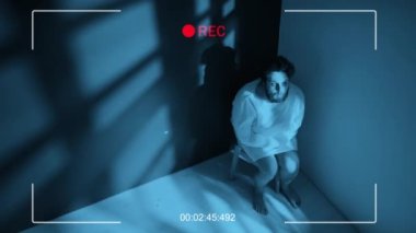 Yüksek açılı CCTV 'de, şizofreni hastası deli gömleği giymiş, koğuşun köşesinde oturan suratını buruşturan, çığlık atan ve kafasını sallayan genç bir adam görülüyor.
