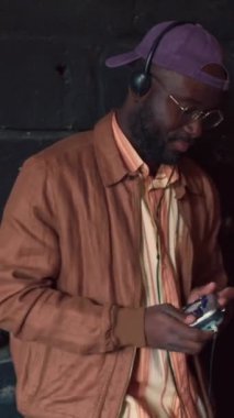 Gündelik kıyafet giyen, kişisel stereo çalar ve kulaklık kullanan koyu gri duvar arkaplanlı, müzik dinleyen genç Afrikalı bir adamın dikey orta portresi.