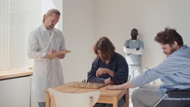精神障害を持つ若者は突然チェスゲームを中断し 攻撃的な方法で仲間の患者を迷惑にし 医療従事者は彼を連れ去った — ストック動画