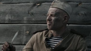 Yüzünde toz ve çamur olan olgun Sovyet askerinin orta boy portresi siperlerde oturup savaş bıçağının keskinliğini kontrol ediyor.