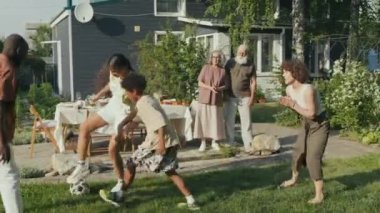 Etnik çeşitlilikte bir aile güneşli bir yaz gününde arka bahçede futbol oynamaktan zevk alıyor, dedem ve ninem maç izliyor.