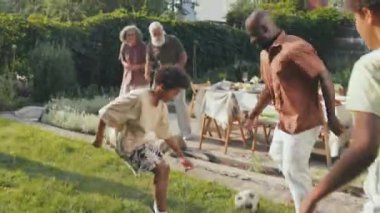 Güneşli bir yaz gününde arka bahçede futbol oynayan modern etnik çeşitlilikte bir ailenin el kamerası.