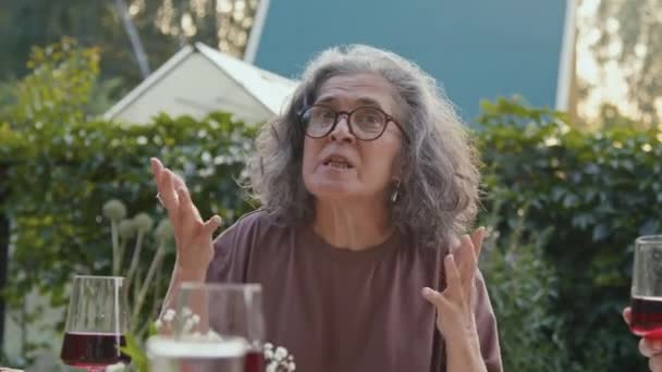 家族の夕食の間に面白い話を伝える裏庭のテーブルにカーリーヘアが座っている現代のおばあちゃんのミディアムクローズアップポートレート — ストック動画