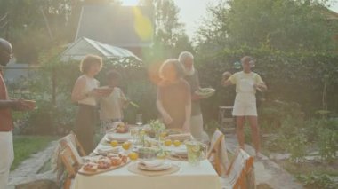 Çocuklar sabun köpüğüyle oynarken, etnik çeşitlilikte aile üyeleri arka bahçeye yemek masası kuruyor.
