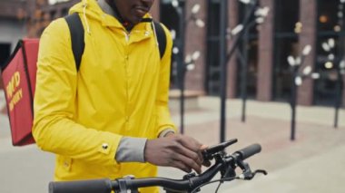 Erkek kuryenin telefonunun orta kısmı bisikletle bağlanıyor. Sarı ceket, beyaz kask ve sırt çantası gibi teslimat ekipmanları giyiyor.
