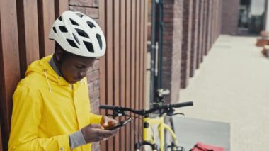 Orta boy yemek dağıtım elemanı duvara yaslanmış, mesaj atıyor, yemek siparişini bekliyor. Bisikleti ve çantası yanında.