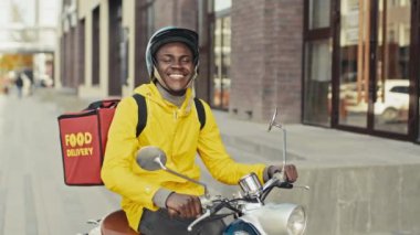 Scooter 'da oturan, sarı ceketli ve mavi kasklı bir teslimatçının portresi. Şehir merkezinde bir yerde kameraya mutlu bir şekilde bakıyor.