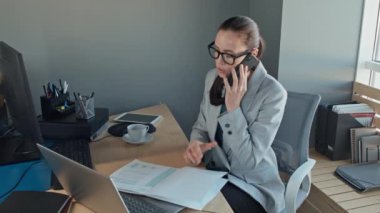 Beyaz kadın iş kadınının telefon görüşmelerine cevap verirken mali raporları gözden geçirirken ve dizüstü bilgisayarda yazarken orta yavaş çekimi.