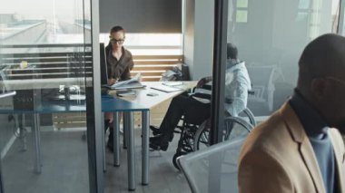 Ofiste dizüstü bilgisayarda çalışan siyah erkek çalışanın orta boy, beyaz kadın yönetmenin arka plandaki tekerlekli sandalyedeki başka bir çalışana iş meselelerini açıklarken görüntüsü.