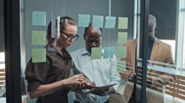 Çok ırklı iş adamlarının orta boy yavaş çekim görüntüleri. Kafkas kadın çalışanı pencereye etiket yapıştırıyor ve siyah erkek iş arkadaşları tablet ve telefon kullanıyor.