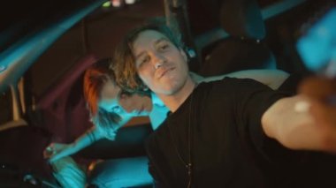 Z Gen beyaz çifti arabada otururken ve park yerinde telefonla selfie çekerken orta ölçekli bir fotoğraf.