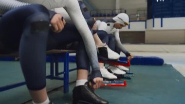 Düşük açılı, çok ırklı buz pateni takımının profesyonel paten takımlarıyla el kamerasıyla kayarken, bağcıklarını bağlarken ve buz pateni sahasında buzun üzerinde yürümeye hazırlanırken.