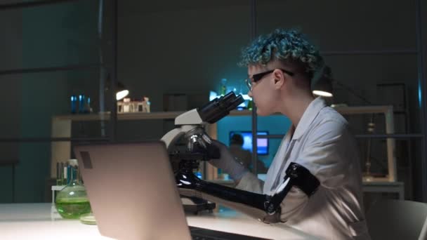 中弧线拍摄的年轻白种人女技师与假臂 白色外套和手套单独在化学实验室工作 在显微镜下观察样品 然后在笔记本电脑上打字 — 图库视频影像