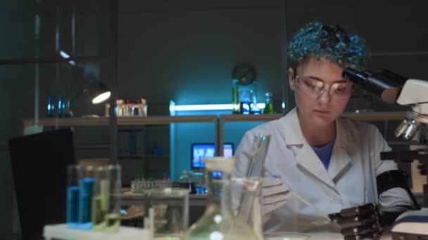 白种人年轻女研究科学家 身穿白衣 坐在生化实验室 将化学溶液倒在培养皿上 显微镜下观察 — 图库视频影像