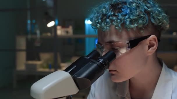 青いカーリーの髪を持つ若い女性の白人研究者の顔のクローズアップショット 薄暗く点灯した生化学実験室で単独で作業し 顕微鏡を調べながら実験を行っている — ストック動画
