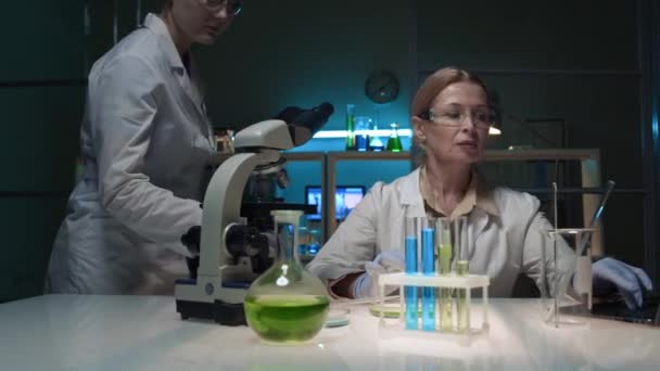 在生物化学实验室工作的中年女性研究人员 在笔记本电脑上打字 带着假臂的助手 带着装有绿色液体的瓶子 有说有笑 — 图库视频影像
