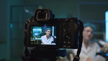 Beyaz önlüklü ve gözlüklü genç kadın bilim adamını kamerayla yakından çekerek laboratuvarda masasında otururken, parlak kimyasal çözümlerle mataraları gösterirken çevrimiçi konferans verirken,