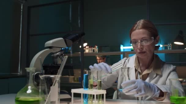 中年の白いコートとガラスで白いコートと青と緑の溶液をフラスコに注ぐ白いコートとメガネの女性研究科学者の中型ショット それをかき混ぜ 反応を見て ラップトップにタイピング — ストック動画