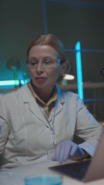 白いコート 手袋および化学実験室で働くメガネの中年白いコートの研究者の中年の縦のショット バイアルの青い解決を調べ そしてラップトップ コンピュータでタイプして下さい — ストック動画