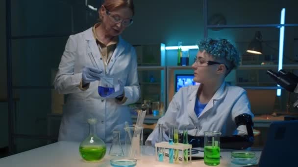 中等比例的残疾女科学家坐在化学实验室 中年同事走进来 展示蓝色液体瓶 与绿色 对话和写作相比较 发表在期刊上 — 图库视频影像