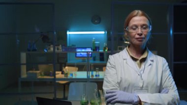 Arka planda katlanmış kollar, şişeler ve mikroskopla kameraya bakarken, biyokimya laboratuvarında poz veren kendinden emin orta yaşlı bir kadının orta boy portresi.