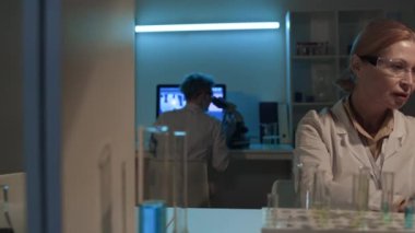 Protez kollu genç bir kadın araştırmacının elektronik mikroskopla hücre kültürüne bakarken orta boy fotoğrafı, sonra da laptopunda yazı yazan olgun beyaz profesöre şişeyi verirken.