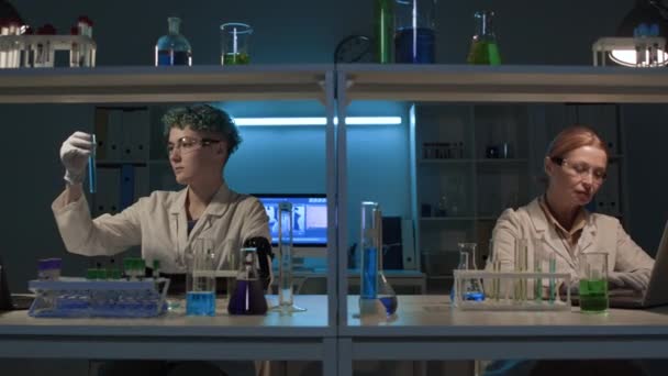 バイアルで青い化学ソリューションを混合する義肢を持つ若い女性技術者の中型ショット その後 成熟したコーカサス科学者にそれを与え ラップトップでタイピングデスクに座って 研究を行っている間 — ストック動画