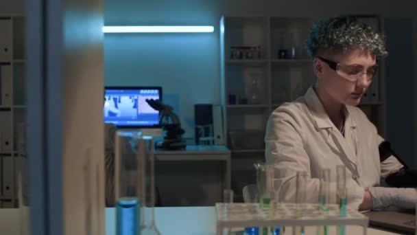 生化学研究所で働く2人の科学者 ラップトップで人工腕タイピングレポートを持つ若い女性 電子顕微鏡で細胞培養を見ている成熟した女性の同僚のミディアムショット — ストック動画