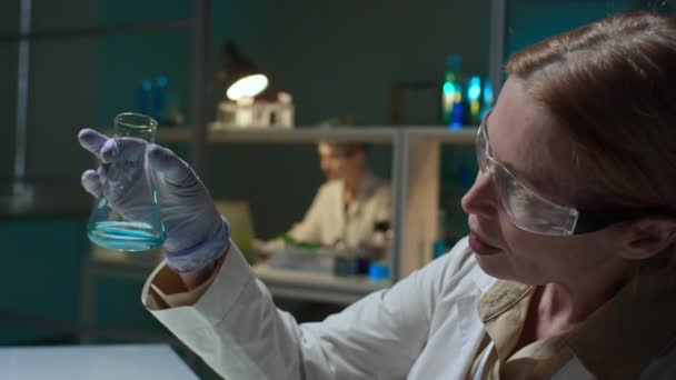 成熟した白人女性科学者の顔と手が青い化学ソリューションでフラスコを見つめ ラップトップでレポートを埋め 生化学研究所で働いている間 — ストック動画