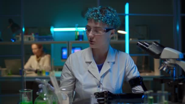 白いコートの若い白いコートの白い女性研究者の中型ショット 実験室の机に座って人工的な腕 緑の化学液体のフラスコを示し オンラインブログのための彼女の仕事について話す — ストック動画