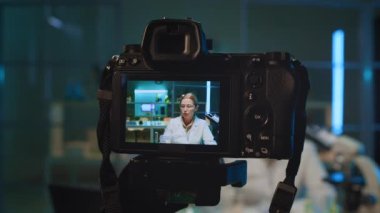 Yetişkin kadın kimyagerin laboratuarda oturup deney yaptığı videoyu kameraya yakın çekimde, mavi sıvıyla matarayı gösterirken çevrimiçi blog için içerik kaydederken,