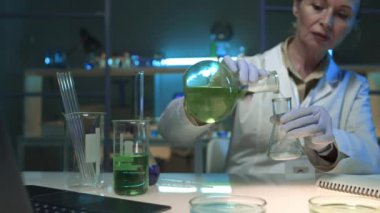 Beyaz yaban domuzu içinde, biyokimya laboratuvarında çalışan, cam kavanozdan mataraya yeşil madde döken, sonra kameraya bakıp gülümseyen olgun beyaz kadın araştırmacının orta boy portresi.